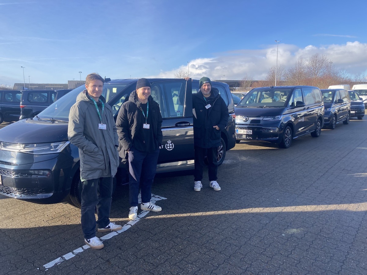 Tre Gire-chauffører med navneskilte i et stort parkeringsområde ved siden af en række mørke Volkswagen varebiler